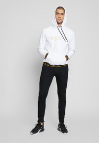 Sweatshirt branca com logotipo dourado, cordões pretos e bolso frontal. Combinado com jeans pretos e sapatilhas de desporto pretas. Detalhe da cintura elástica visível.