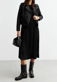 Musta nahkainen moottoripyörätakki mustan mekon päällä, yhdistettynä mustiin nilkkabootseihin, joissa on hopeisia yksityiskohtia. Kantaa pientä mustaa käsilaukkua.