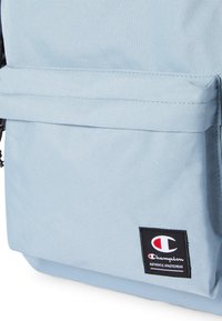 Bolsa de tela azul claro con un bolsillo, que presenta un parche del logo de Champion en negro con una "C" roja y blanca y el texto "Authentic Athleticwear".