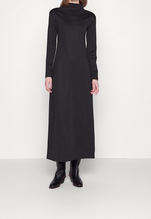 Frau trägt ein langes schwarzes Kleid mit hohem Kragen und langen Ärmeln sowie schwarze Knöchelstiefel, steht vor einem schlichten hellen Hintergrund.