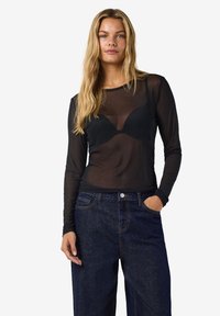 Ženska z dolgimi blond lasmi, oblečena v prozorno črno dolgo majico preko črnega nedrčka in temno modrih kavbojkah, stoji z eno roko v žepu.