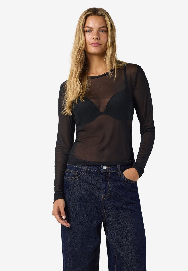 Ženska z dolgimi blond lasmi, oblečena v prozorno črno dolgo majico preko črnega nedrčka in temno modrih kavbojkah, stoji z eno roko v žepu.