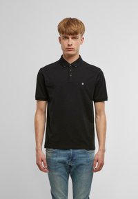 Fekete poloing klasszikus gallérral és három gombbal. Rövid ujjú, a mellkasán egy kisebb logó található. Kék farmernadrággal viselve.