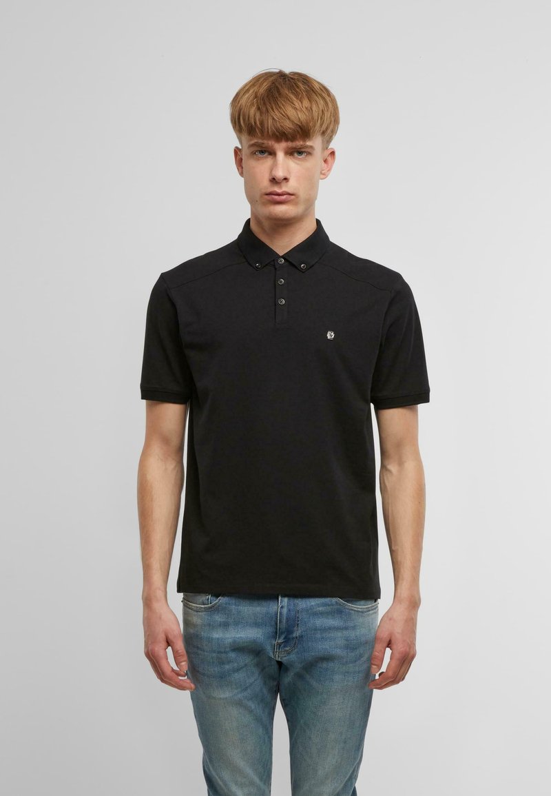 Fekete poloing klasszikus gallérral és három gombbal. Rövid ujjú, a mellkasán egy kisebb logó található. Kék farmernadrággal viselve.