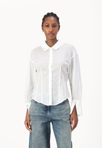 Chemise blanche à boutons avec col, fabriquée en tissu léger. Elle présente des manches longues, un ourlet arrondi et de subtils détails brodés sur les manches.