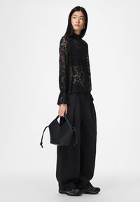 Top de renda preta de manga comprida com punhos franzidos, combinada com calças largas pretas. A segurar uma pequena mala preta estruturada.