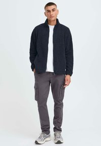 Giacca zip-up plush blu scuro, t-shirt bianca, pantaloni cargo grigi con tasche laterali e sneakers grigie. La giacca ha una finitura morbida e testurizzata.