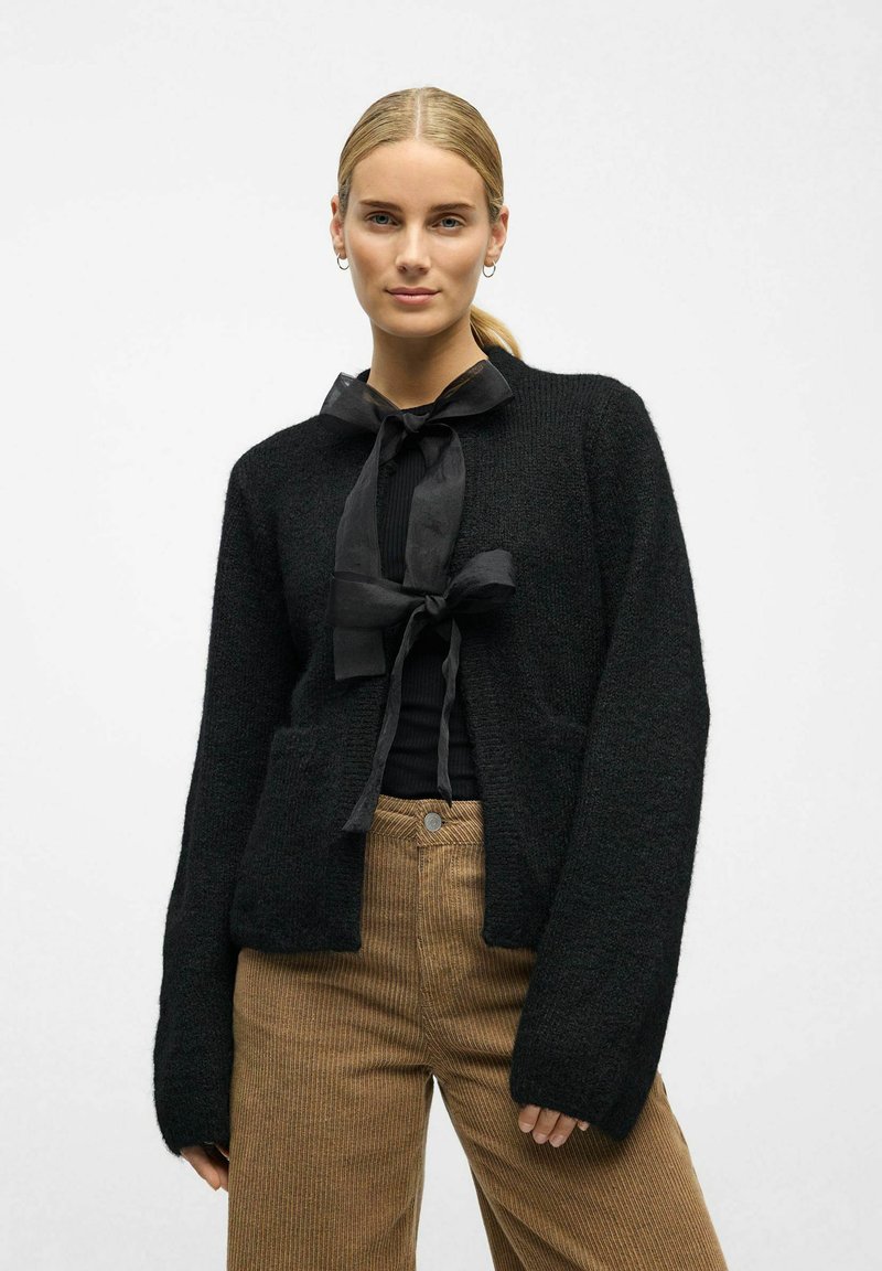 Černý pletený kardigán s volným střihem, dlouhými rukávy a předními kapsami. Má černou stuhu na zavazování u límce, v kombinaci s ribbovanými béžovými kalhotami.