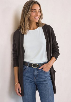 Vrouw met lichtbruin haar draagt wit overhemd, donker vest, blauwe spijkerbroek en zwarte riem, lacht en kijkt naar haar rechterkant tegen een effen achtergrond.