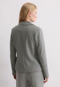 Blazer gris à fines rayures avec un col structuré, une coupe ajustée et une texture lisse, porté sur une tenue grise assortie.