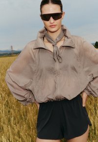 Femme portant une veste beige oversize, un short noir, une écharpe grise autour du cou et de grandes lunettes de soleil noires, debout dans un champ de blé.