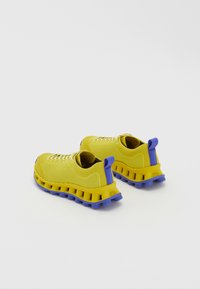 Κίτρινα πλεκτά αθλητικά παπούτσια με μπλε ελαστικά στοιχεία και μαλακή σόλα, που διαθέτουν θηλιά στο πίσω μέρος και κορδόνια για ασφαλή εφαρμογή.