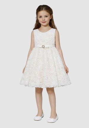 Giovane ragazza in piedi, che indossa un vestito bianco senza maniche con trama floreale e cintura, abbinato a ballerine bianche, su uno sfondo neutro.