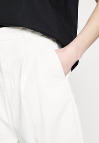 Personne portant un pantalon blanc avec la main placée dans la poche avant et portant un haut noir.
