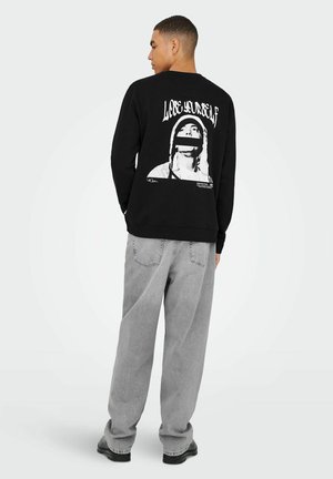 Schwarzes Langarmshirt mit einem großen weißen Grafikmotiv auf dem Rücken, auf dem der Text "Lose Yourself" steht, kombiniert mit hellgrauen, weiten Hosen.
