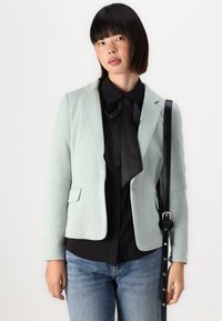 Světlý mátově zelený blazer s upraveným střihem, s klopami a dvěma předními kapsami, v kombinaci s černou blůzkou a džínovými kalhotami. Viditelný černý ramenní popruh.