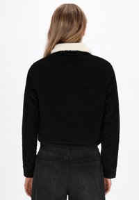 Mustat corduroy-takit, joissa on kermaisen vaaleat karvakaulus, lyhyt leikkaus ja teksturoitu kangas. Sopii käytettäväksi rentoihin tilaisuuksiin, pitkät hihat ja takana hihna.