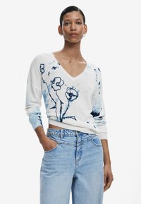 Weißer V-Ausschnitt-Pullover mit blauem Blumenmuster, langen Ärmeln und gerippten Manschetten. Kombiniert mit hellblauen, hochtaillierten Jeans.