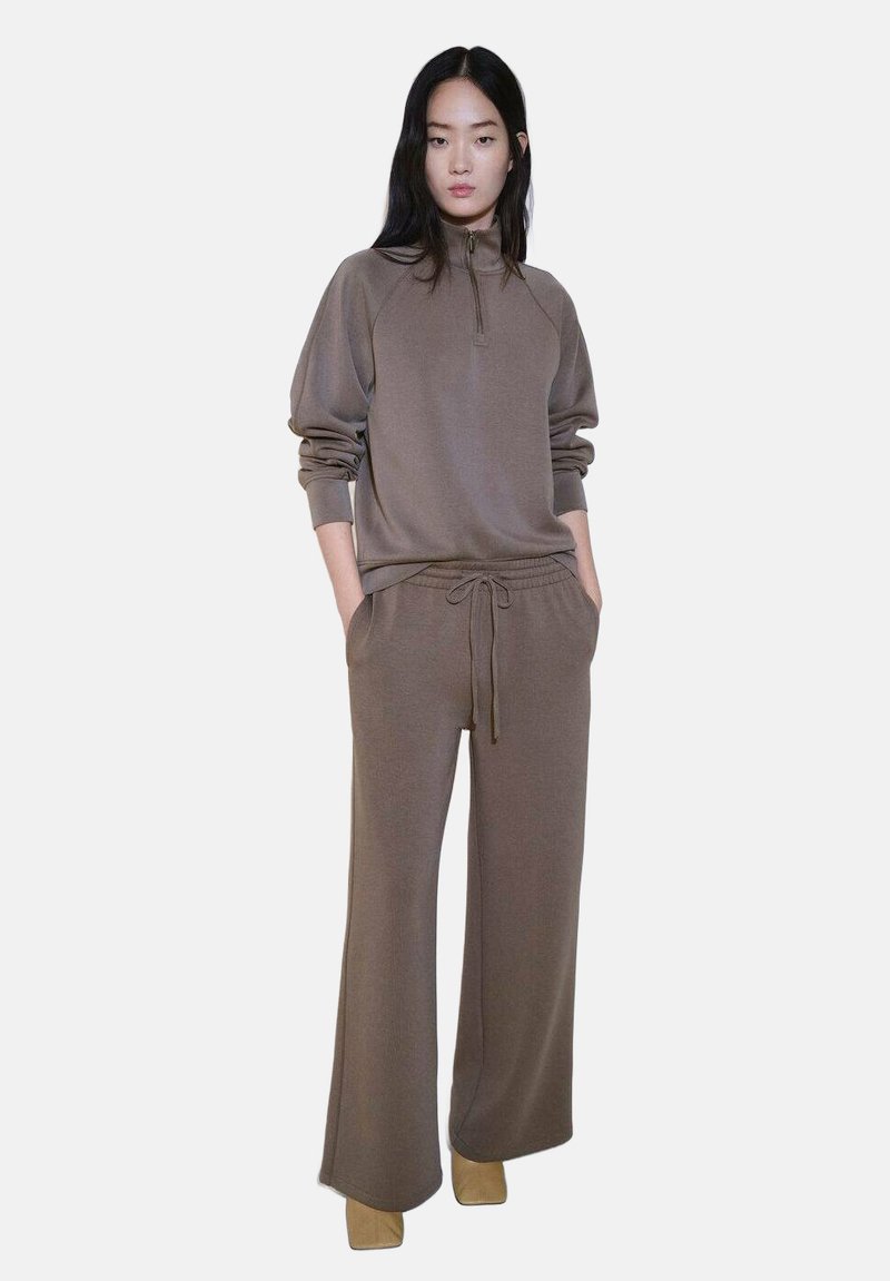 Smeđa dukserica s polu-zip ovratom u kombinaciji s odgovarajućim hlačama širokih nogavica, oboje napravljeno od mekog materijala. Model stoji s rukama u džepovima.