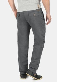 Pantalon gris léger avec poches arrière fermées par des boutons, coupe droite et ajustement décontracté. Porté avec des chaussures sombres sans lacets.