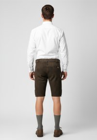 Braune Shorts aus strukturiertem Stoff, mit einer Reißverschlusstasche und dekorativen Knöpfen an den Seiten. Kombiniert mit einem weißen Langarmshirt.