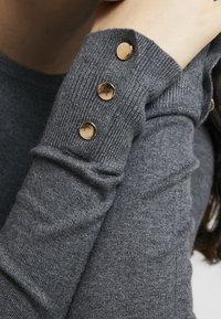 Détail d'un pull en maille grise avec des poignets côtelés et trois boutons ronds dorés sur un côté pour l'accent. Texture douce et coupe ajustée.