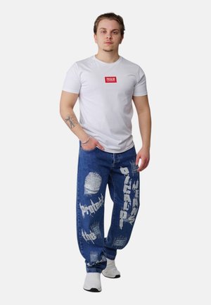 Junger Mann steht, trägt ein weißes T-Shirt mit rotem Logo, blaue Jeans mit weißem grafischem Text und Aufnähern, weiße Turnschuhe und ein Tattoo auf dem linken Unterarm.