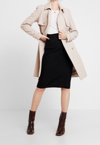 Beige trenchcoat yllä valkoista ribbattua toppia ja mustaa ribbattua kynähametta, yhdistettynä ruskeisiin korkokenkiin nilkkojen ylle. Huomio keskittyy siistiin, rakenteelliseen muotoiluun.