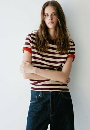 Jeune femme aux longs cheveux bruns portant un t-shirt rayé à manches courtes et un jean bleu foncé, debout les bras croisés devant un fond uni.