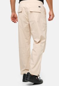Beige cargo bukser med en afslappet pasform, der har sidelommer og en glat tekstur. Båret med sorte sneakers og synlig elastik ved anklerne.