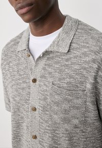 Grijze gebreide poloshirt met een textuurpatroon, korte mouwen, een kraag en een borstzak; heeft bruine knopen aan de voorkant.