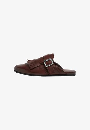 Zapato marrón de cuero tipo slip-on con detalle de flecos, perforaciones decorativas y un acento de hebilla plateada. Presenta una suela plana negra.