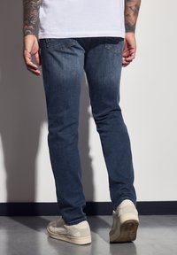 Mörkblå denimjeans med slim fit, som har subtila blekningar, bakfickor och slitna vita sneakers. Slät textur.