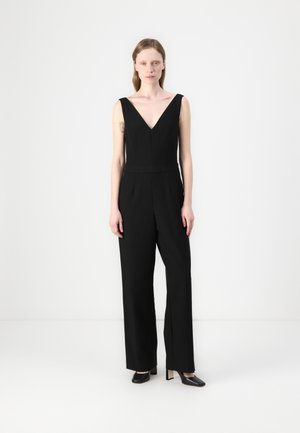 Zwart jumpsuit met een diepe V-halslijn, wijde pijpen en gestructureerde naden. Gecombineerd met zwarte hoge hakken. Effen achtergrond.