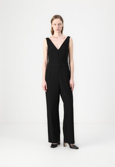 Mono negro con escote en V profundo, diseño de pierna ancha y costuras estructuradas. Combinado con zapatos de tacón alto negros. Fondo liso.