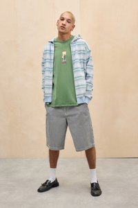 Young man with short blond hair wearing green graphic t-shirt, striped zip-up hoodie, black-and-white checkered shorts, white socks, and black shoes.