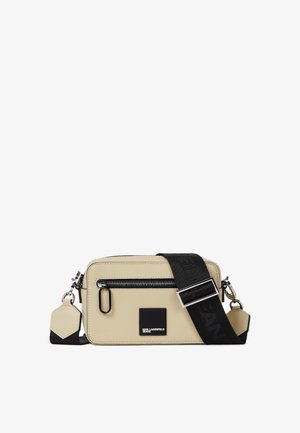 Sac bandoulière rectangulaire beige avec fermeture éclair noire, patch logo noir et large sangle noire réglable avec boucle argentée.