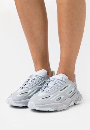 Beneden aan het lichaam met lichtgrijze sportsneakers met meshpanelen, vetersluiting en dikke, gestructureerde zolen op een witte achtergrond.