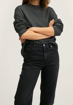 Personne portant une chemise à manches longues gris foncé avec les manches retroussées et un jean noir taille haute, les bras croisés sur la poitrine.