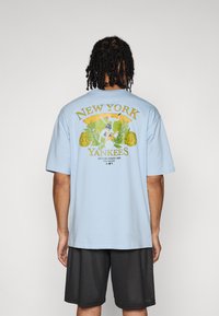 T-shirt en coton bleu clair avec un graphique coloré des New York Yankees au dos, incluant des images de baseball et un feuillage décoratif.