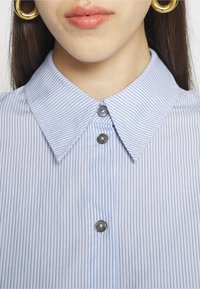 Camicia a righe verticali blu chiaro con colletto a punta, caratterizzata da due bottoni grigi davanti e texture di cotone morbido.