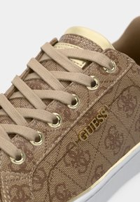 Braune Leinensneaker mit einem sich wiederholenden Logo-Muster, Textillaces, goldfarbigen Ösen und einer weißen Gummisohle.