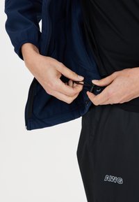 Marinblå zip-jacka med elastiska manschetter, justerbar dragsko och en enfärgad svart skjorta under. Mörka byxor med "AWG"-logotyp.