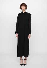 Vestido negro de manga larga con cuello tipo camisa, con pliegues en el frente y una silueta fluida, combinado con tacones negros de tira en el tobillo.