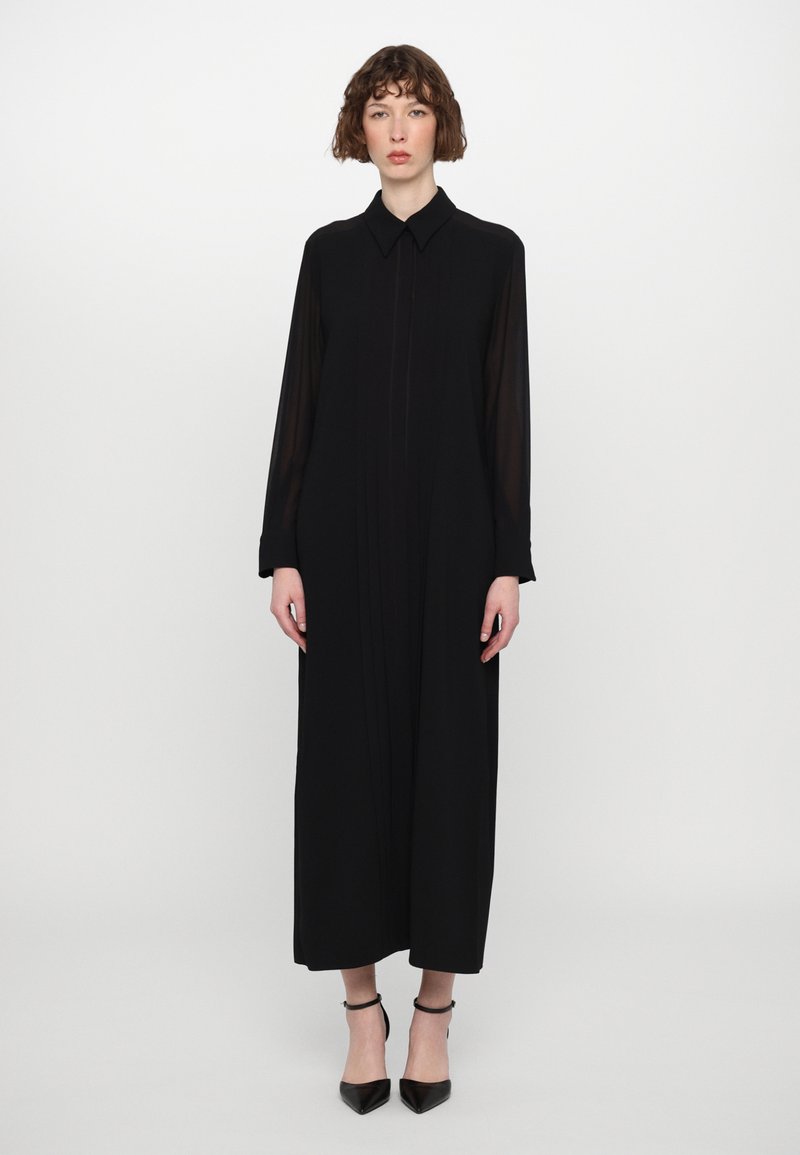 Vestido negro de manga larga con cuello tipo camisa, con pliegues en el frente y una silueta fluida, combinado con tacones negros de tira en el tobillo.