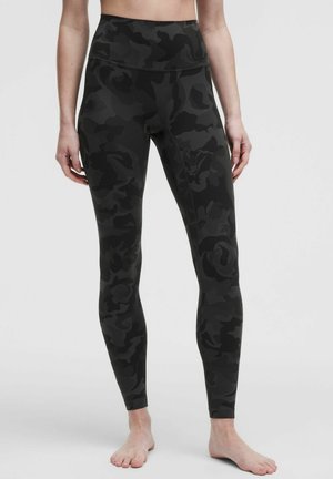 Kobieta nosząca czarne i ciemnoszare legginsy z wysokim stanem i wzorem moro, stojąca boso na jednolitym tle.