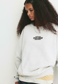 Felpa grigia con la scritta "GROWN WITH FEELINGS". Presenta una vestibilità ampia, polsini a costine e un look a strati con una striscia gialla sotto.