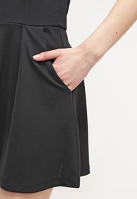 Czarna sukienka wykonana z gładkiej tkaniny, z rozkloszowanym dołem, marszczeniami i boczną kieszenią z ręką wsuniętą w środku.