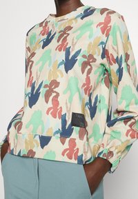 Sweat-shirt en coton multicolore avec des motifs abstraits de feuilles en vert, bleu, beige et bordeaux, doté d'un col rond côtelé et de poignets élastiques.