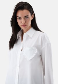White button-up shirt featuring a large heart pocket with ruffle detailing on the left side. Smooth, lightweight fabric, relaxed fit.
